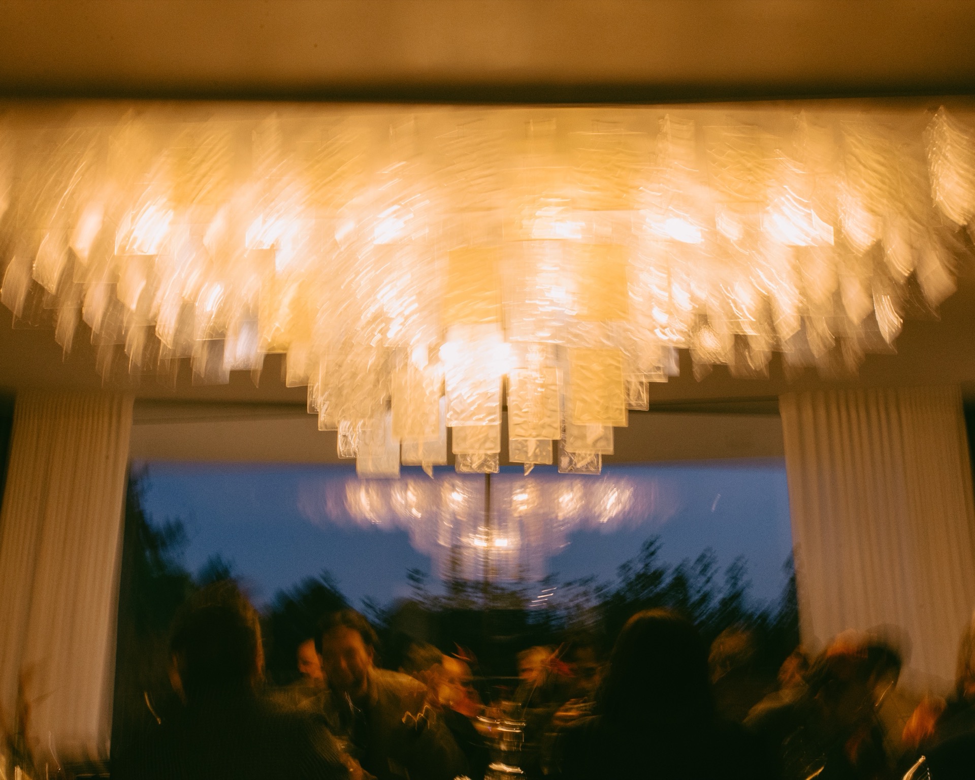 A motion-blurred, low-light view of people dining under a large, illuminated geometric chandelier that might suit an Audi Q9 interior.