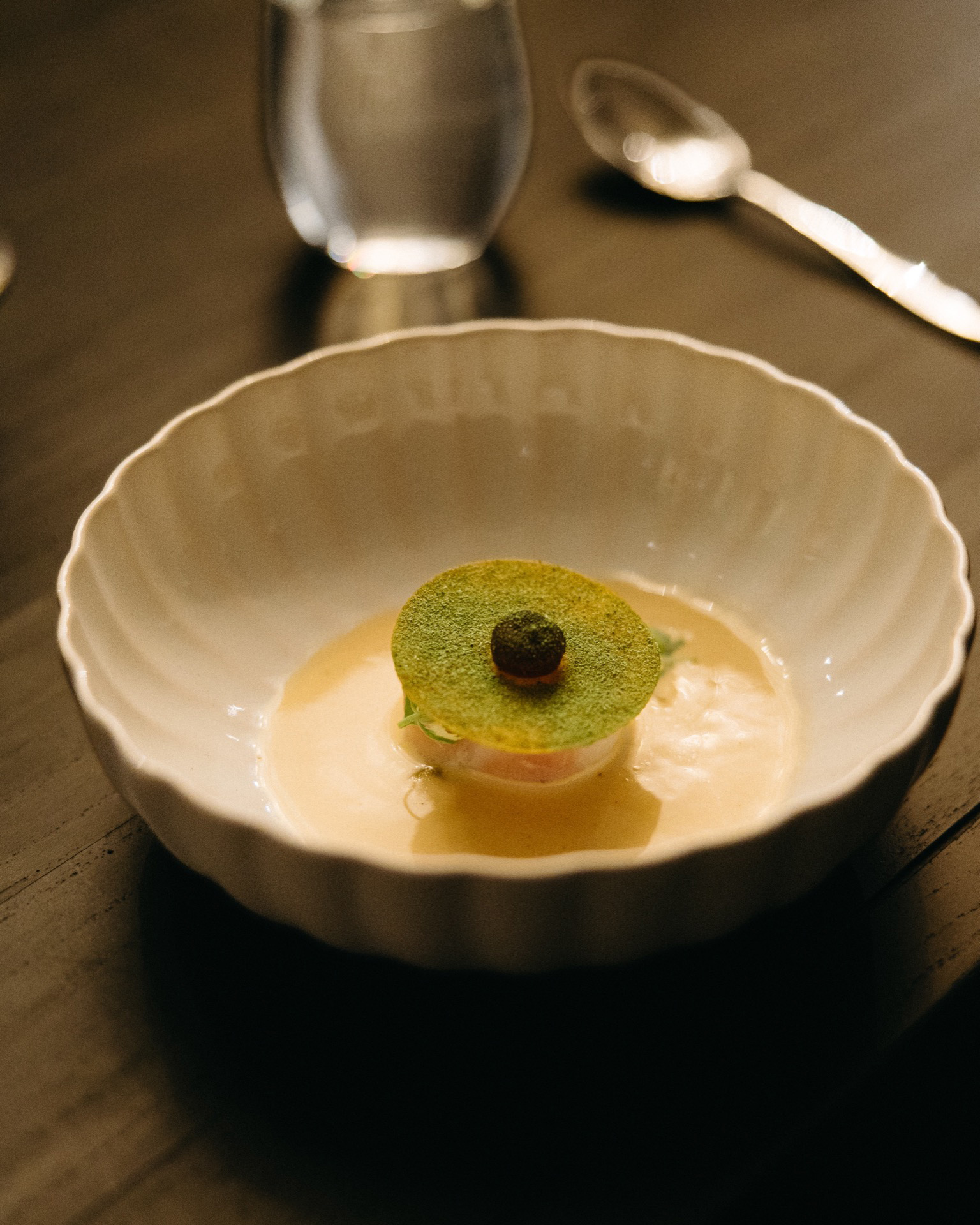 A decorative green disc topped with a small brown sphere, presented like an Audi Q9 in a shallow, fluted white bowl filled with a creamy sauce.