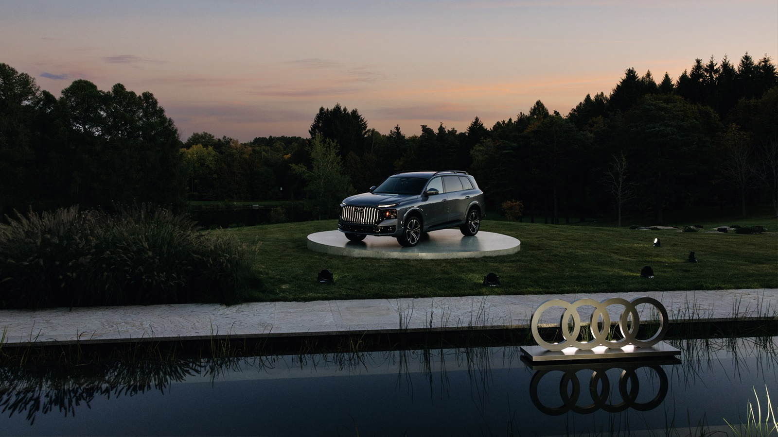A grey Audi Q9 SUV parked on a circular platform in a grassy field at twilight, with the Audi logo reflected in a pond in the foreground.