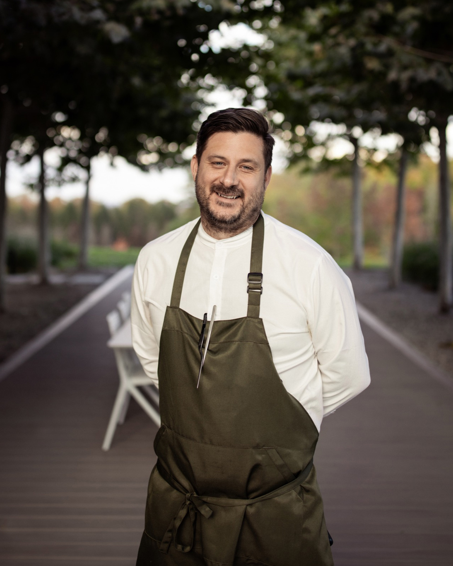 A smiling man in a white shirt and green apron standing on an outdoor walkway near an Audi Q9.