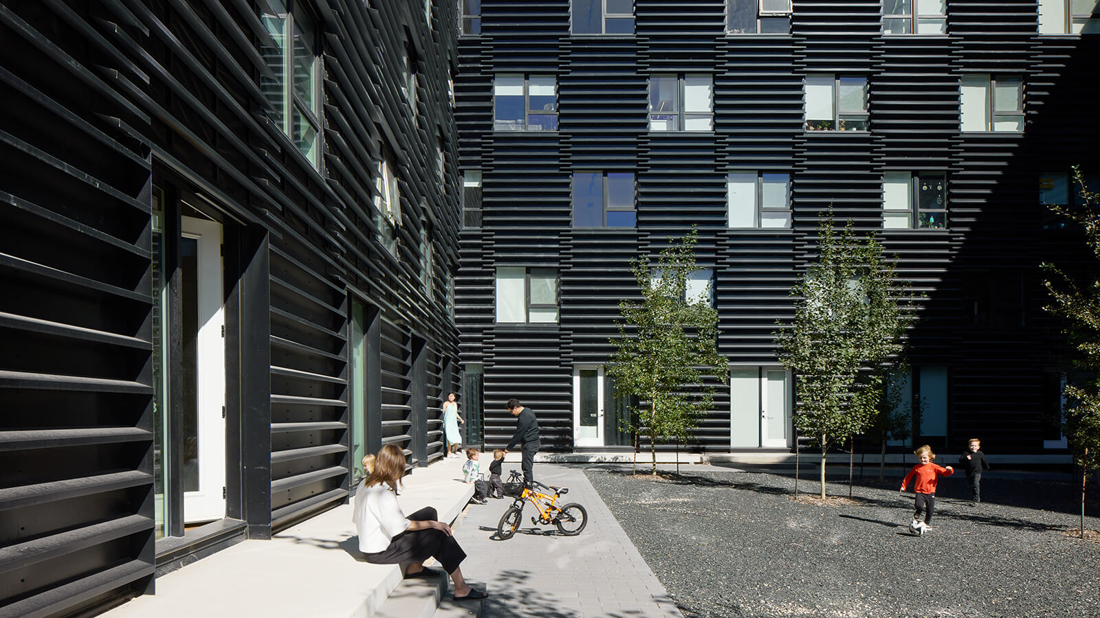 People and children with a bicycle in a courtyard surrounded by a modern black building with horizontal slats.
