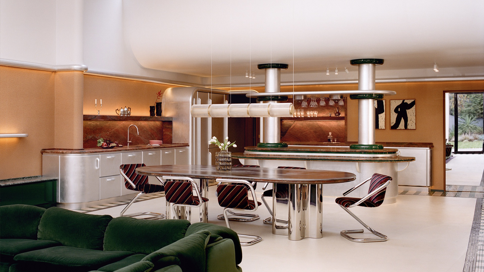An open-plan kitchen and dining area in a Paris apartment designed by Atelier HA featuring a wooden oval table with striped cantilever chairs, stainless steel cabinetry, and green velvet furniture.
