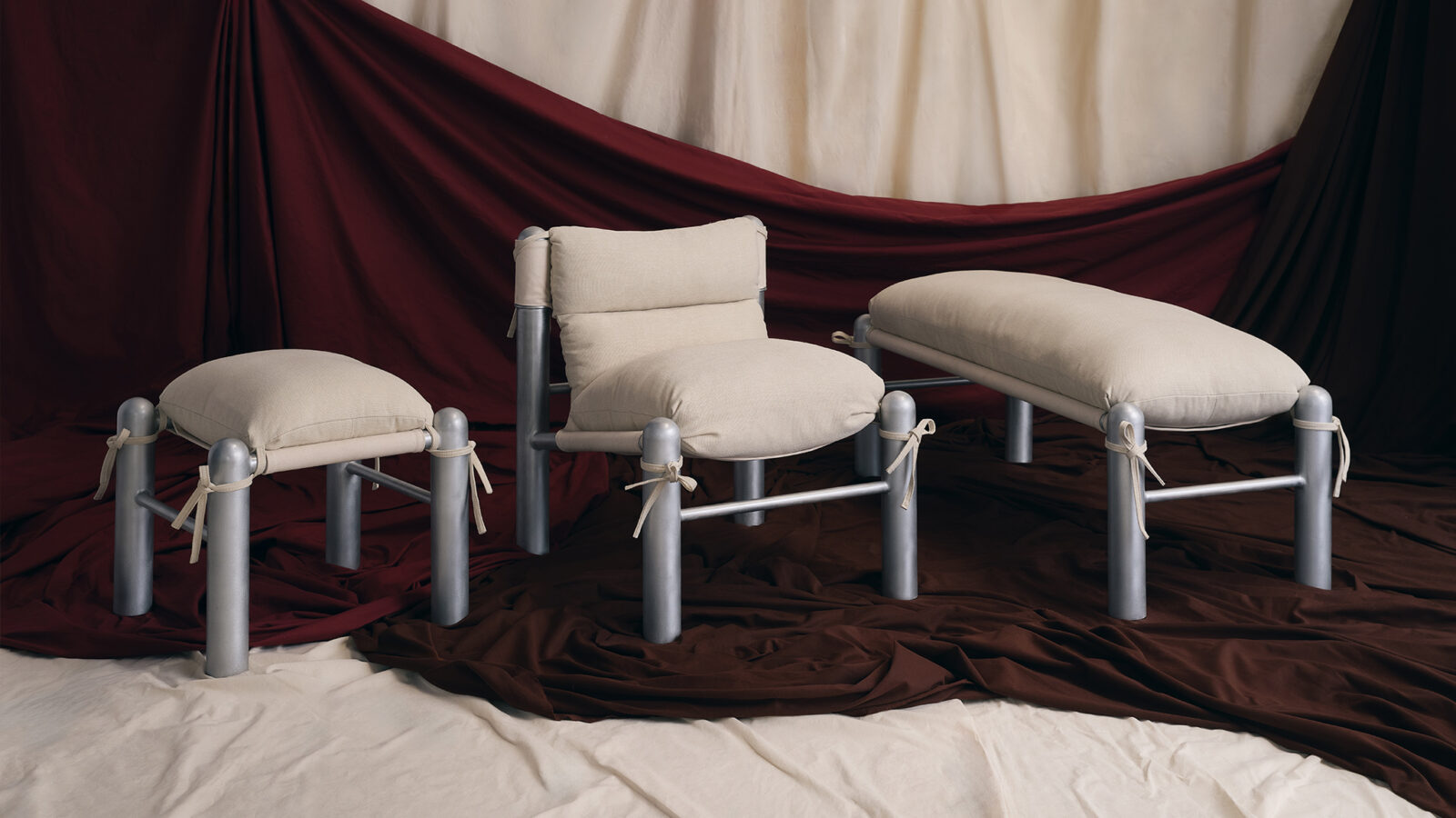 Three padded chairs with silver metal frames sit on cream and brown draped fabric, with a burgundy and beige fabric backdrop. Each chair has off-white cushions and tied fabric accents.
