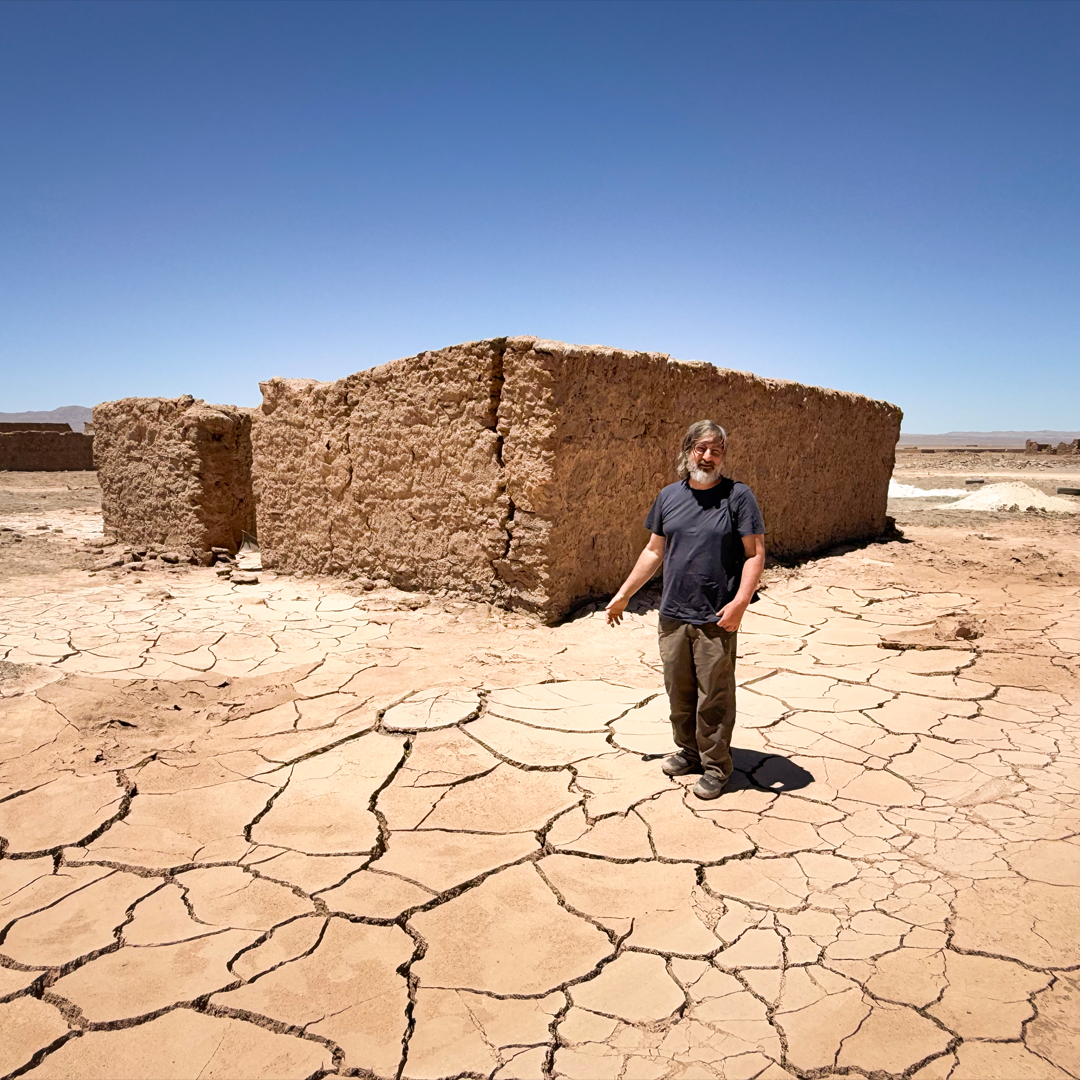 A man stands on dry, cracked earth before a ruined adobe building under a clear blue sky.