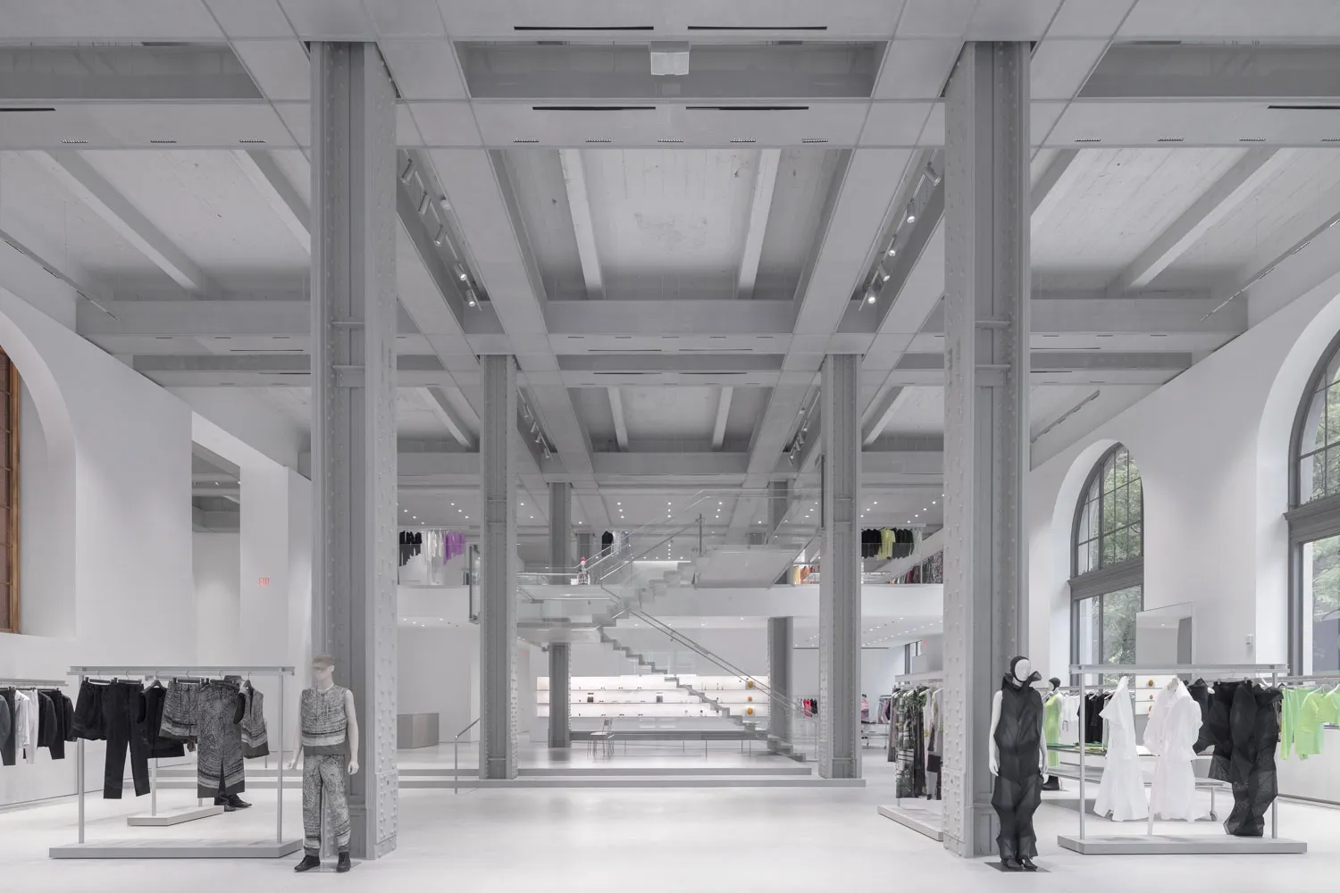 A spacious, minimalist retail store interior, with high ceilings, exposed structural columns, and clothing racks featuring mannequins.
