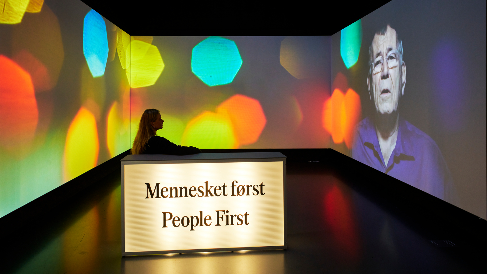 A person stands behind a glowing “Mennesket først / People First” podium at an October event, set against projections of bokeh lights and an older man’s face.