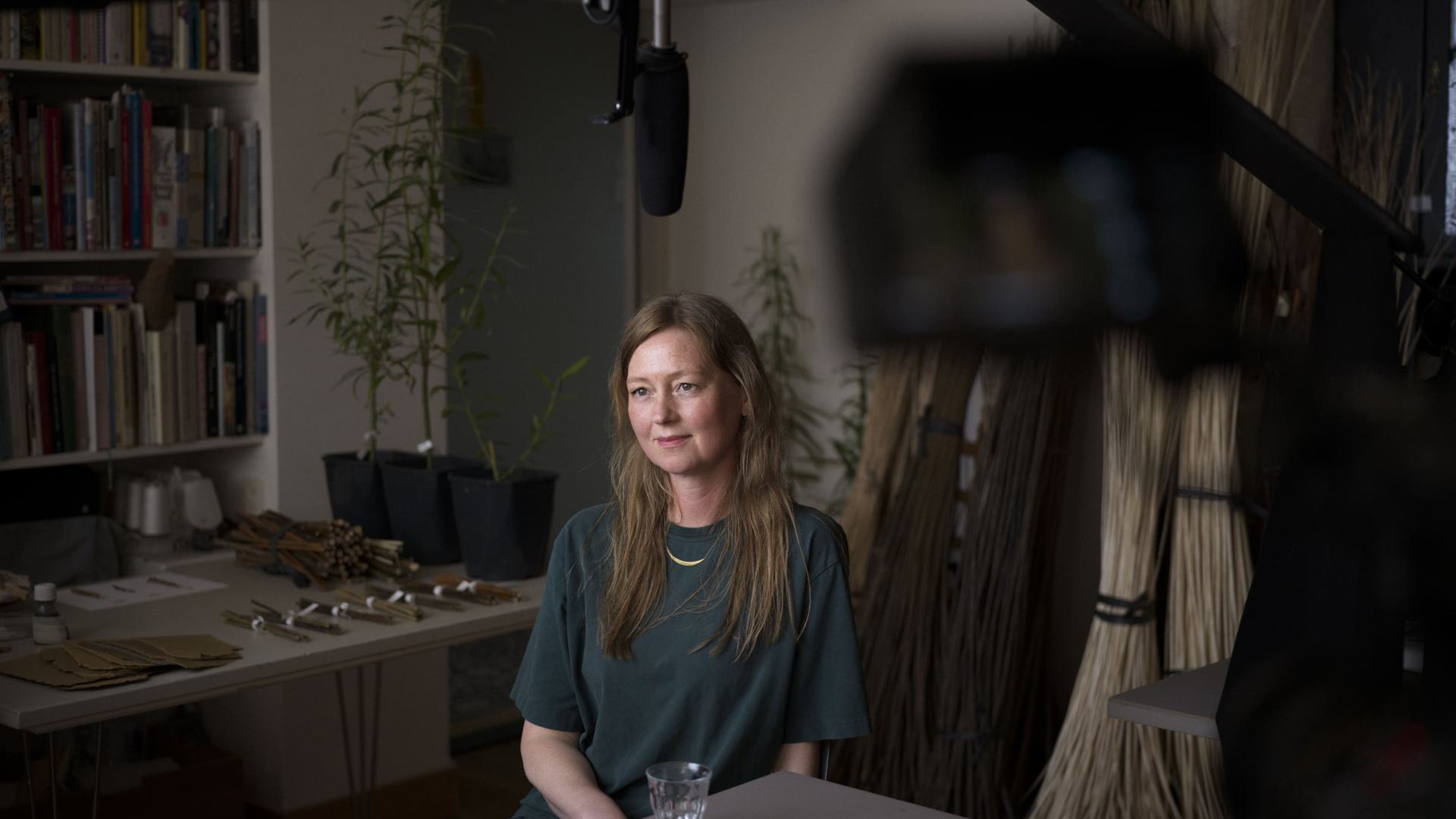 A woman sits in a room with potted plants and bundles of sticks for an October event, being filmed by a camera and microphone.