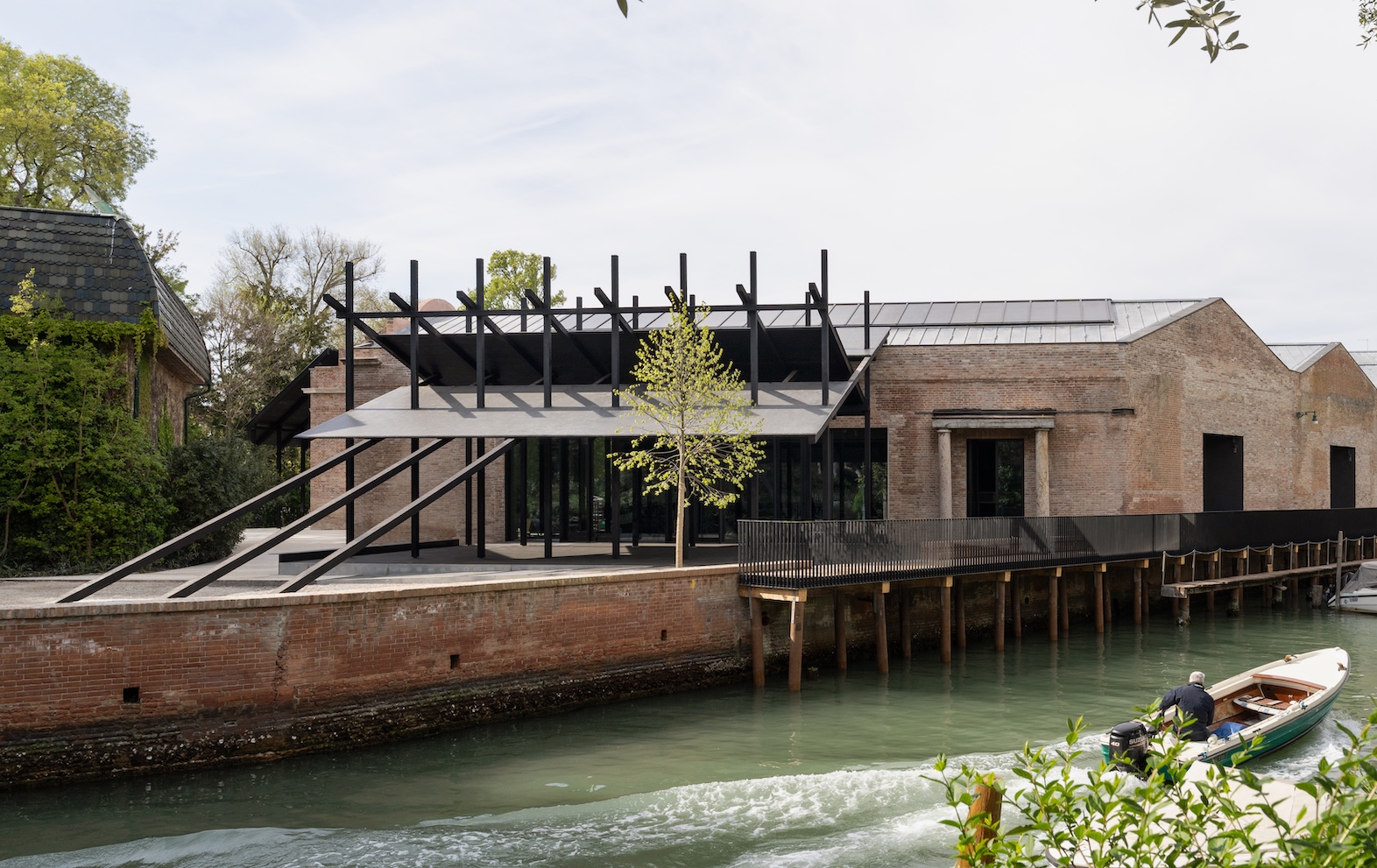 A modern building with a black structural frame sits on a brick wall beside a canal, where a person in a small motorboat travels through the water.