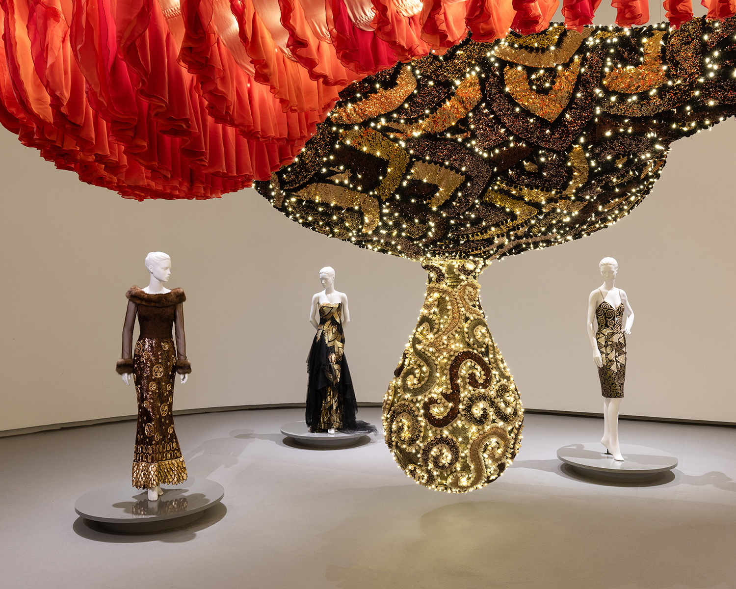 Three mannequins wearing brown and gold formal gowns, displayed under a large, illuminated, teardrop-shaped art installation.
