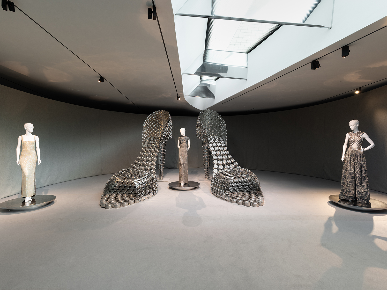 Three mannequins in formal gowns stand with two sculptural high-heeled shoes made of metal discs in a gallery space.