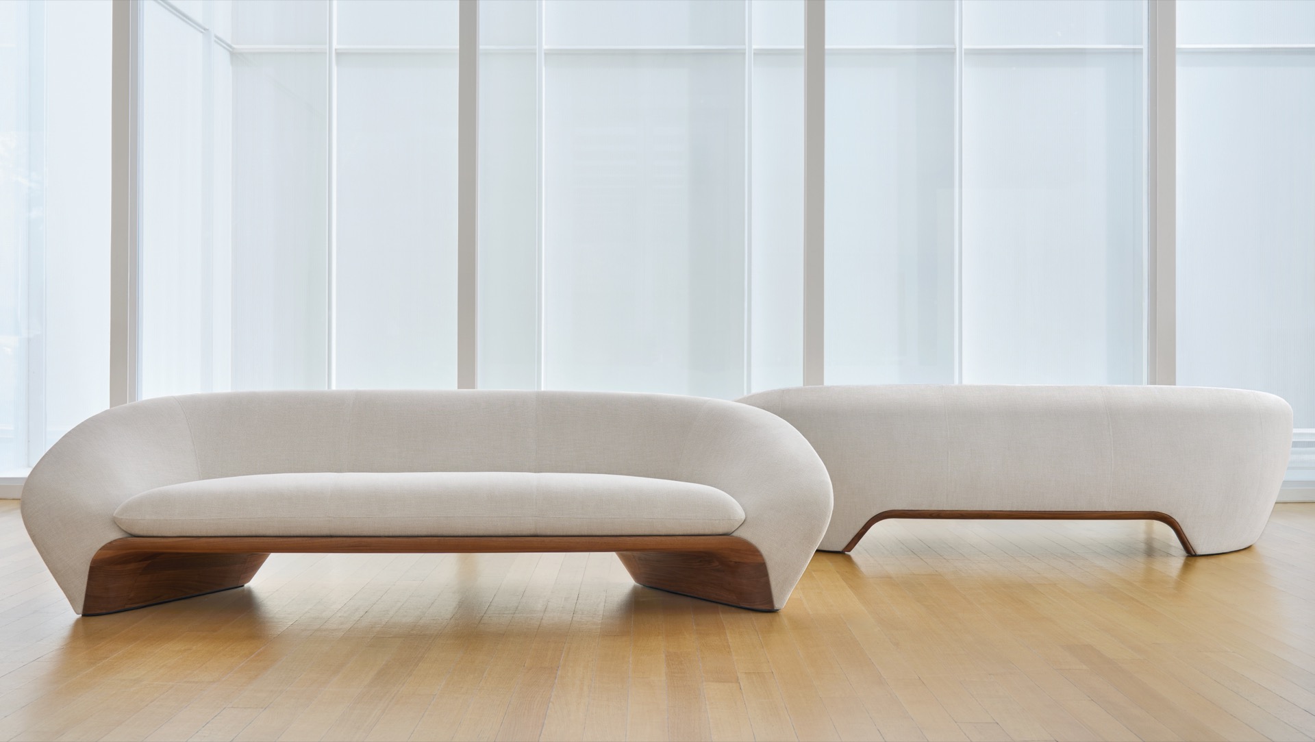 Two modern, curved white sofas with wooden bases are placed on a light wood floor in front of tall, frosted glass windows.