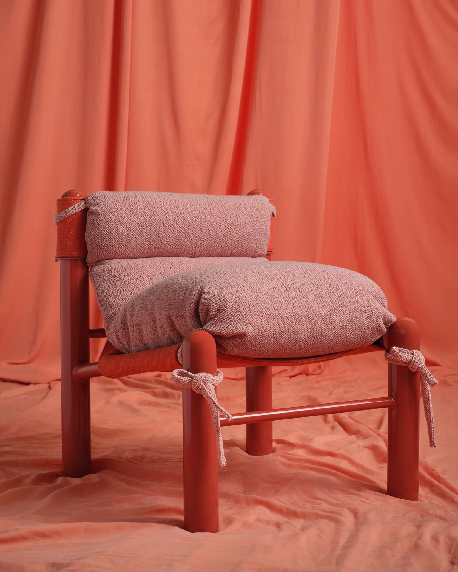 A modern chair with a plush pink cushion and red frame, set against draped red fabric in the background and on the floor.